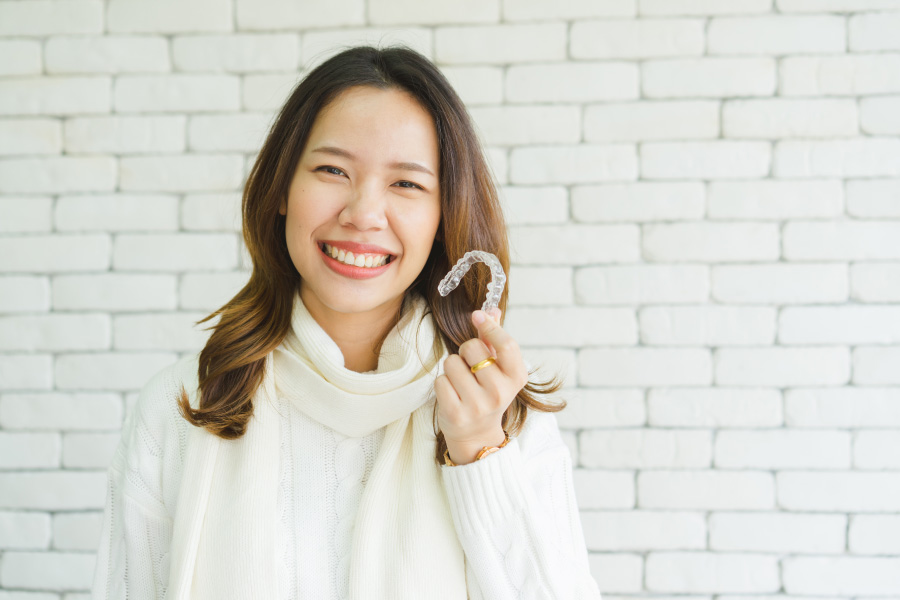 Asian woman in a cream sweater smiles against a white brick wall while holding Invisalign clear aligners