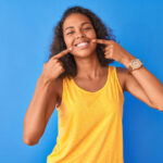 Brown woman in a yellow tank top points at her smile after improving it with cosmetic dentistry