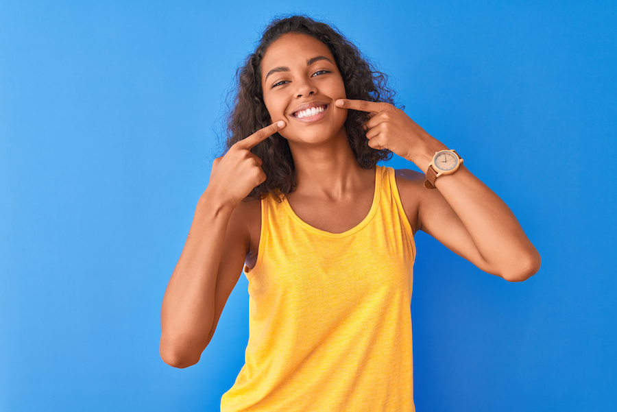 Brown woman in a yellow tank top points at her smile after improving it with cosmetic dentistry, dentist in Hot Springs Village